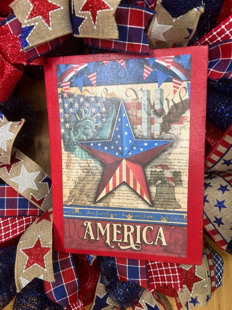 May include: A rectangular wooden sign with a red border featuring a central star with American flag patterns, the Statue of Liberty, a bald eagle, and the Liberty Bell. The text AMERICA is printed at the bottom, surrounded by patriotic ribbon.