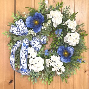 May include: A circular wreath featuring green foliage, white hydrangea clusters, and large blue poppy-like flowers. A decorative ribbon with a blue and white floral pattern and striped edges is tied to the side, with long tails hanging down the left side.