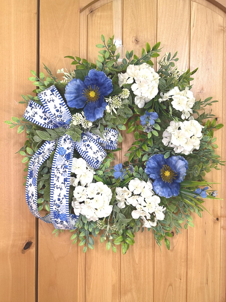 May include: A circular wreath featuring green foliage, white hydrangea clusters, and large blue poppy-like flowers. A decorative ribbon with a blue and white floral pattern and striped edges is tied to the side, with long tails hanging down the left side.