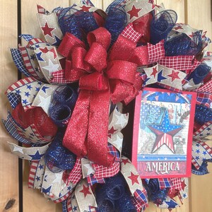 May include: A patriotic wreath featuring red, white, and blue mesh ribbons, star-patterned burlap, and a large red glittery bow. A rectangular sign attached to the right side displays a star graphic and the word AMERICA in white lettering.
