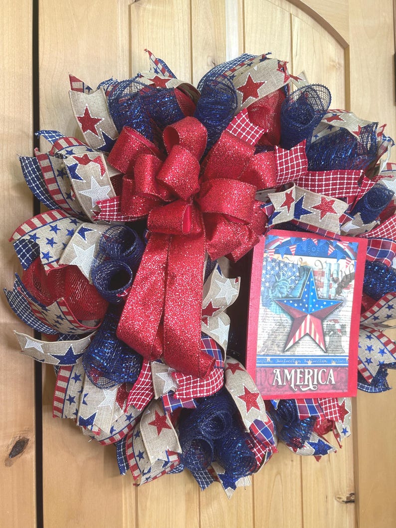 May include: A patriotic wreath featuring red, white, and blue mesh ribbons, star-patterned burlap, and a large red glittery bow. A rectangular sign attached to the right side displays a star graphic and the word AMERICA in white lettering.