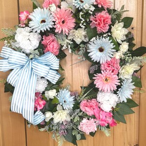 May include: A circular floral wreath featuring a mix of light blue, pink, and white artificial flowers, including daisies and carnations, accented with green leaves and small white filler flowers, finished with a large light blue and white striped bow.