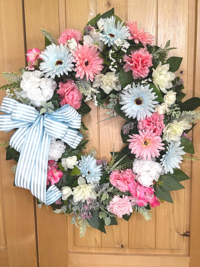 May include: A circular floral wreath featuring a mix of light blue, pink, and white artificial flowers, including daisies and carnations, accented with green leaves and small white filler flowers, finished with a large light blue and white striped bow.
