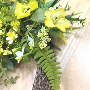 May include: A decorative wreath featuring a circular brown woven twig base adorned with bright yellow artificial flowers, small white blossoms, and various green foliage including fern-like leaves and slender grass-like stems.