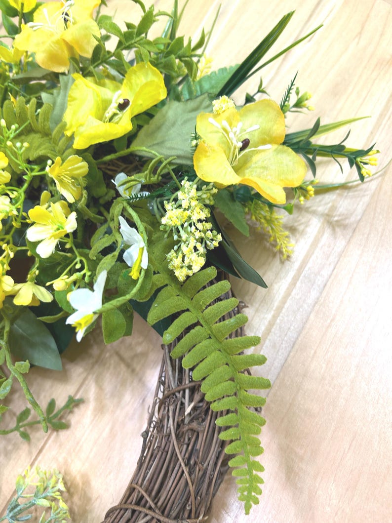 May include: A decorative wreath featuring a circular brown woven twig base adorned with bright yellow artificial flowers, small white blossoms, and various green foliage including fern-like leaves and slender grass-like stems.