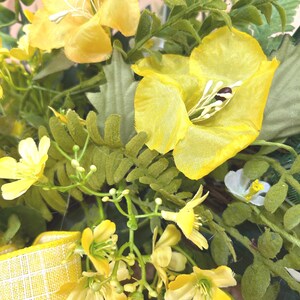 May include: A decorative wreath featuring bright yellow fabric flowers with prominent white stamens, interspersed with various green foliage and fern-like leaves. A yellow and white checkered ribbon is attached to the lower left side.