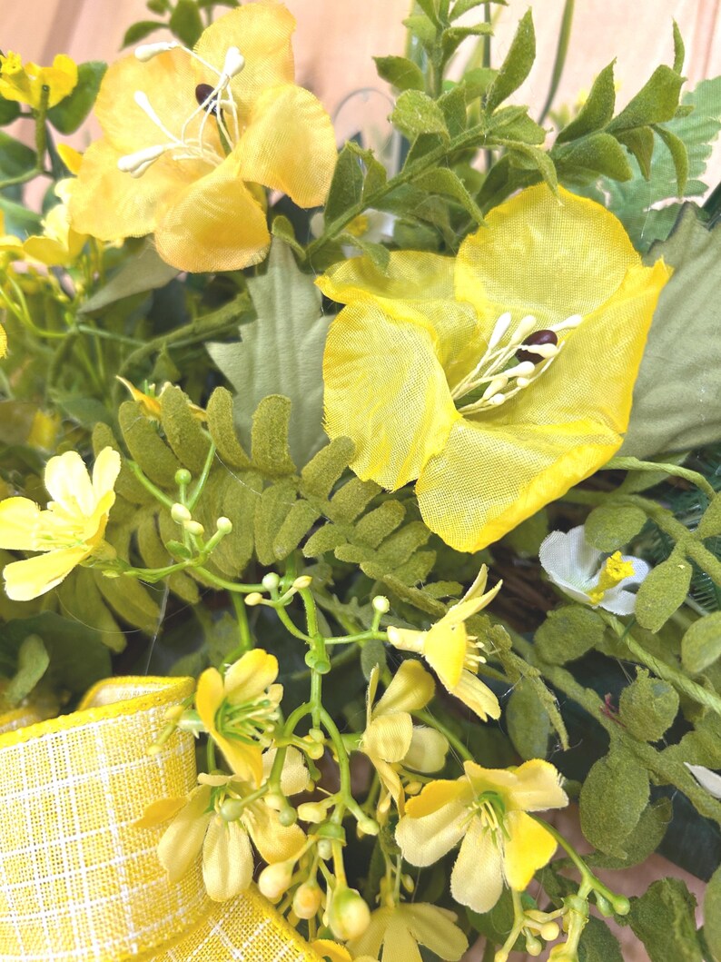 May include: A decorative wreath featuring bright yellow fabric flowers with prominent white stamens, interspersed with various green foliage and fern-like leaves. A yellow and white checkered ribbon is attached to the lower left side.