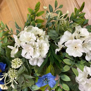 May include: A decorative floral wreath featuring clusters of white hydrangea-like blossoms and smaller white flowers, accented by a single large blue flower with a yellow center, a smaller blue flower, and various green foliage leaves.