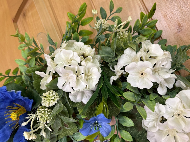May include: A decorative floral wreath featuring clusters of white hydrangea-like blossoms and smaller white flowers, accented by a single large blue flower with a yellow center, a smaller blue flower, and various green foliage leaves.