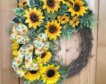 May include: A decorative wreath featuring bright yellow sunflowers with dark brown centers, interspersed with green foliage and small white berries. A patterned ribbon with a sunflower print is tied to the left side of the brown twig base.