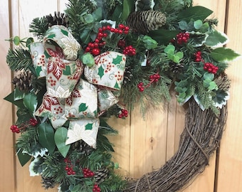 May include: A holiday wreath featuring a natural grapevine base adorned with artificial pine branches, green leaves, and clusters of red berries. A large decorative bow with a cream, red, and green poinsettia pattern is attached to the side.
