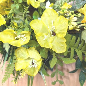 May include: A decorative wreath featuring bright yellow silk flowers with white stamens and dark centers, interspersed with various green foliage including fern-like leaves and small sprigs, creating a dense and layered floral arrangement.