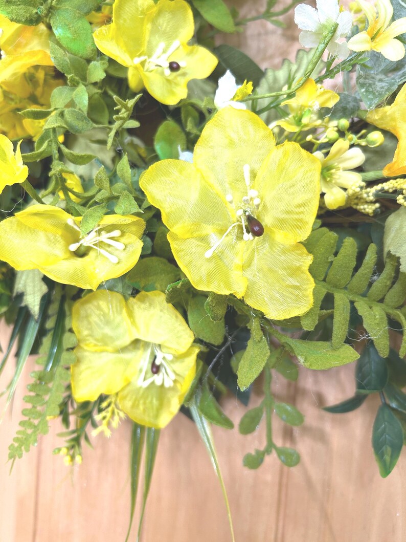 May include: A decorative wreath featuring bright yellow silk flowers with white stamens and dark centers, interspersed with various green foliage including fern-like leaves and small sprigs, creating a dense and layered floral arrangement.