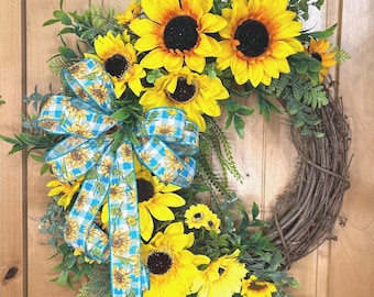 Op de afbeelding: Een decoratieve krans met felgele zonnebloemen met donkere harten, groen blad en een grote strik van blauw-wit geblokt lint met een zonnebloemmotief, alles gerangschikt op een ronde basis van bruine takken.