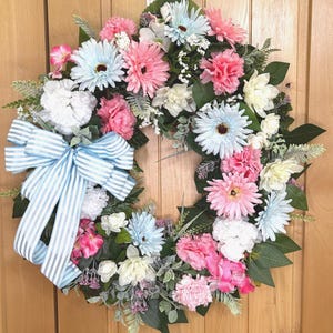 May include: A circular floral wreath featuring a mix of light blue, pink, and white artificial flowers, including daisies and carnations, accented with green foliage and a large, light blue and white striped fabric bow attached to the left side.