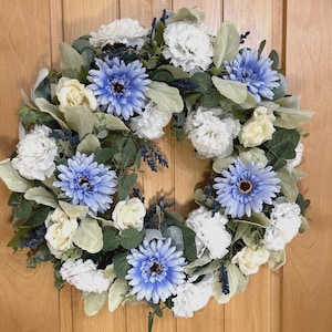 May include: A circular wreath featuring light blue daisy-like flowers, white carnations, and cream-colored roses. The arrangement is accented with soft green lamb's ear leaves, dark green eucalyptus foliage, and small sprigs of dark blue lavender.