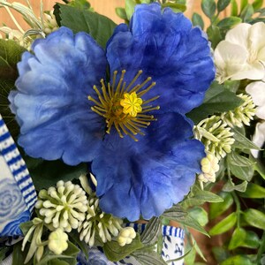 May include: A decorative arrangement featuring a large, vibrant blue flower with a yellow center, surrounded by small white flower clusters and green foliage. A white ribbon with a blue floral pattern is tied at the base of the floral display.