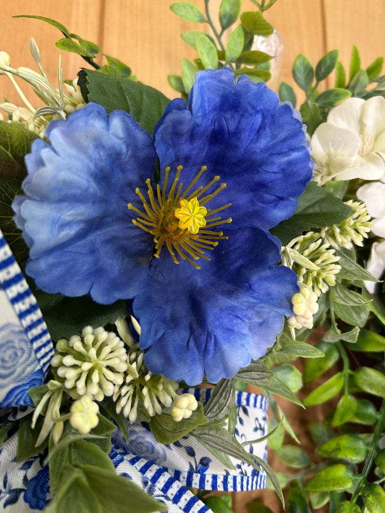 May include: A decorative arrangement featuring a large, vibrant blue flower with a yellow center, surrounded by small white flower clusters and green foliage. A white ribbon with a blue floral pattern is tied at the base of the floral display.