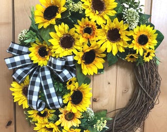 May include: A circular wreath constructed from brown twigs, adorned with numerous bright yellow sunflowers featuring dark brown centers. The arrangement includes green leaves and small white floral clusters, accented by a large black and white buffalo check ribbon bow.