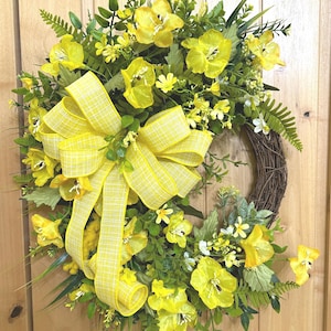 May include: A circular wreath featuring a brown twig base adorned with bright yellow silk flowers, various green foliage, and ferns. A large, prominent yellow and white plaid ribbon bow is attached to the left side, with long streamers hanging down.