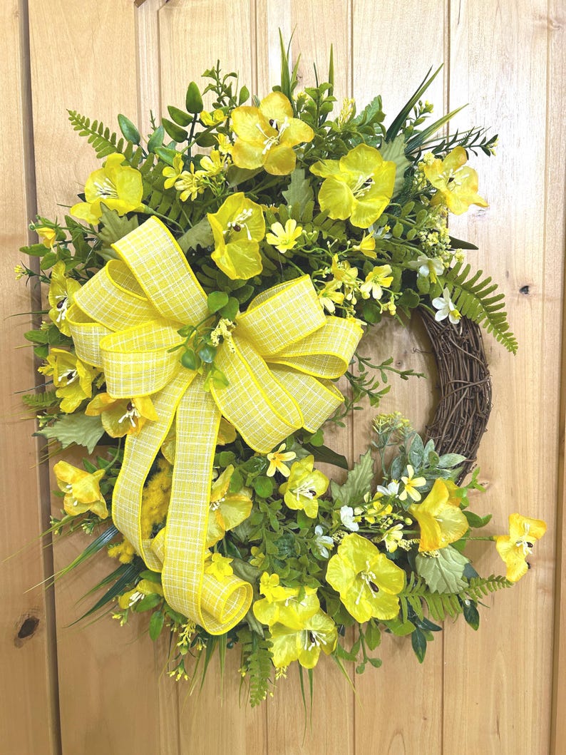 May include: A circular wreath featuring a brown twig base adorned with bright yellow silk flowers, various green foliage, and ferns. A large, prominent yellow and white plaid ribbon bow is attached to the left side, with long streamers hanging down.