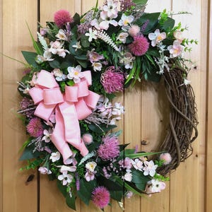 May include: A circular wreath constructed from a natural brown twig base, adorned with an arrangement of green leaves, small white flowers, and vibrant pink thistle-like blooms. A large, multi-looped light pink ribbon is attached to the left side.