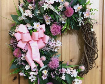 May include: A circular wreath constructed from a natural brown twig base, adorned with an arrangement of green leaves, small white flowers, and vibrant pink thistle-like blooms. A large, multi-looped light pink ribbon is attached to the left side.