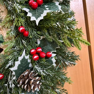 May include: A decorative wreath featuring artificial green cedar branches with a light dusting of white snow. The arrangement includes clusters of bright red berries, green holly leaves with white edges, and brown pinecones throughout the foliage.