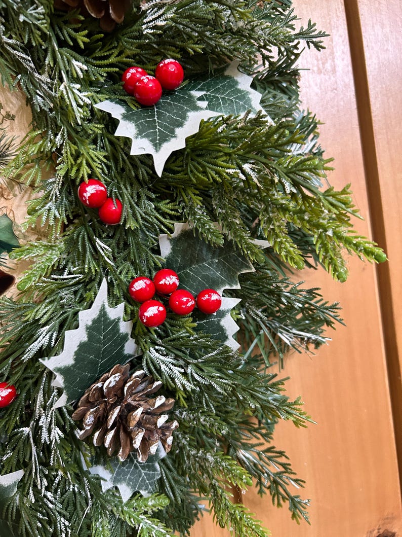 May include: A decorative wreath featuring artificial green cedar branches with a light dusting of white snow. The arrangement includes clusters of bright red berries, green holly leaves with white edges, and brown pinecones throughout the foliage.