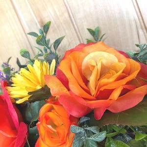 May include: A floral arrangement featuring vibrant orange and red roses, bright yellow daisies, and small purple flower accents. The bouquet includes various green leaves and foliage, creating a dense, colorful display of artificial botanical elements.
