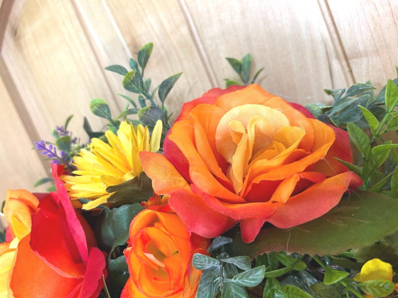 May include: A floral arrangement featuring vibrant orange and red roses, bright yellow daisies, and small purple flower accents. The bouquet includes various green leaves and foliage, creating a dense, colorful display of artificial botanical elements.