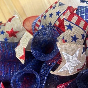May include: A decorative wreath featuring patriotic elements including tan hessian ribbons with red and blue star patterns, red and white striped ribbons, and dark blue metallic mesh rolls. The design incorporates a mix of textures and colours.