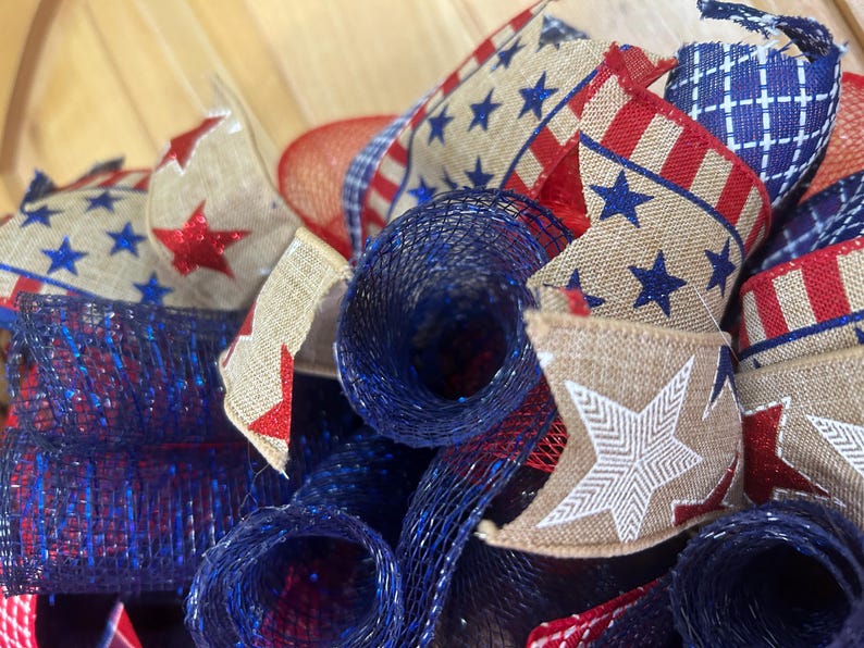 May include: A decorative wreath featuring patriotic elements including tan hessian ribbons with red and blue star patterns, red and white striped ribbons, and dark blue metallic mesh rolls. The design incorporates a mix of textures and colours.