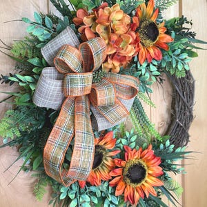 May include: A decorative wreath featuring orange and yellow sunflowers, green foliage, and a central plaid ribbon bow with orange, brown, and white checkered patterns. The wreath is constructed on a circular brown twig base with various green leaves.