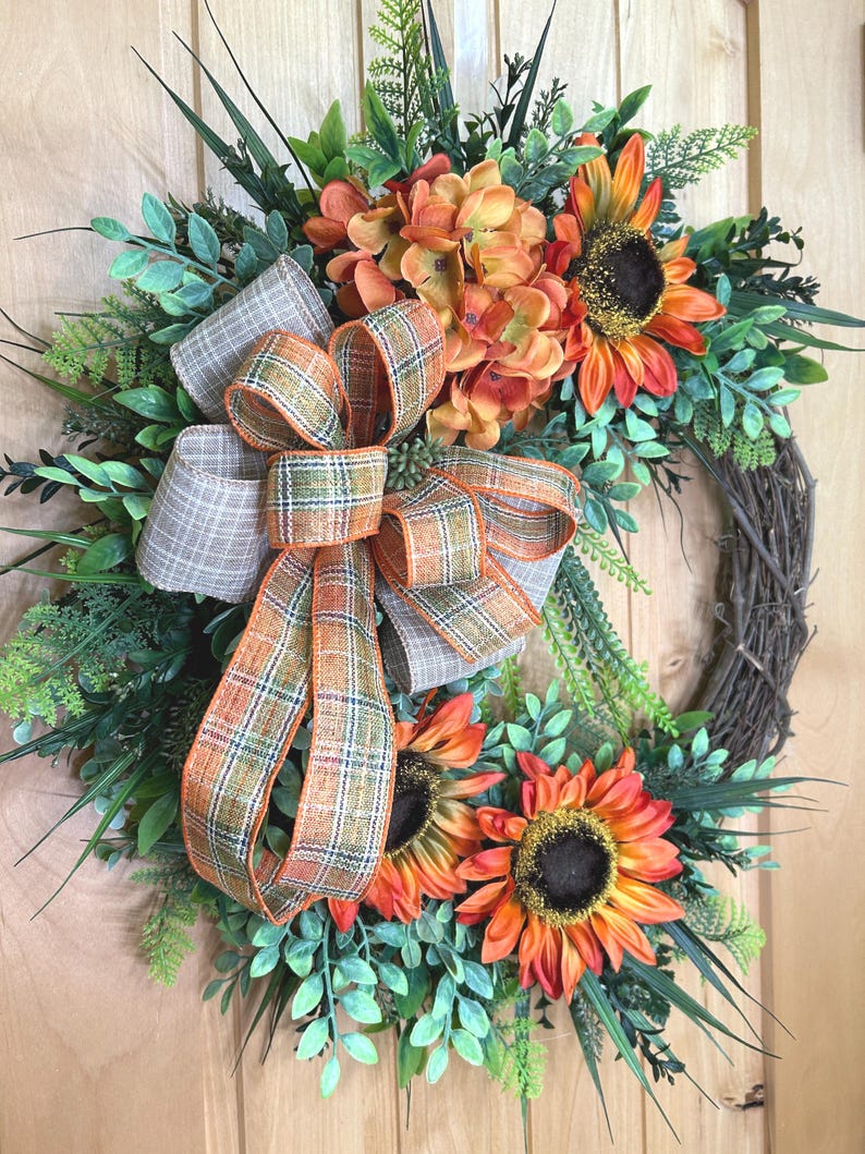 May include: A decorative wreath featuring orange and yellow sunflowers, green foliage, and a central plaid ribbon bow with orange, brown, and white checkered patterns. The wreath is constructed on a circular brown twig base with various green leaves.