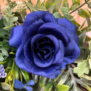 Op de afbeelding: Een diepblauwe kunstmatige roos als middelpunt met gelaagde stoffen bloemblaadjes. De centrale roos is omringd door diverse groene bladeren, waaronder eucalyptusachtige bladeren, kleine witte bloemknoppen en kleine blauwe accentbloemen.