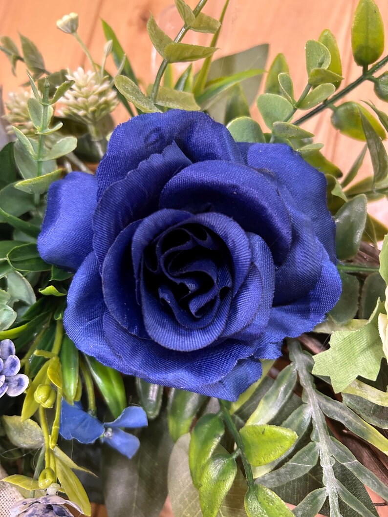 Op de afbeelding: Een diepblauwe kunstmatige roos als middelpunt met gelaagde stoffen bloemblaadjes. De centrale roos is omringd door diverse groene bladeren, waaronder eucalyptusachtige bladeren, kleine witte bloemknoppen en kleine blauwe accentbloemen.