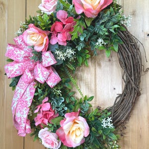 May include: A decorative wreath featuring a natural brown grapevine base adorned with artificial pink and cream roses, vibrant pink hydrangea clusters, and green foliage. A large, patterned pink and white floral ribbon bow is attached to the side.