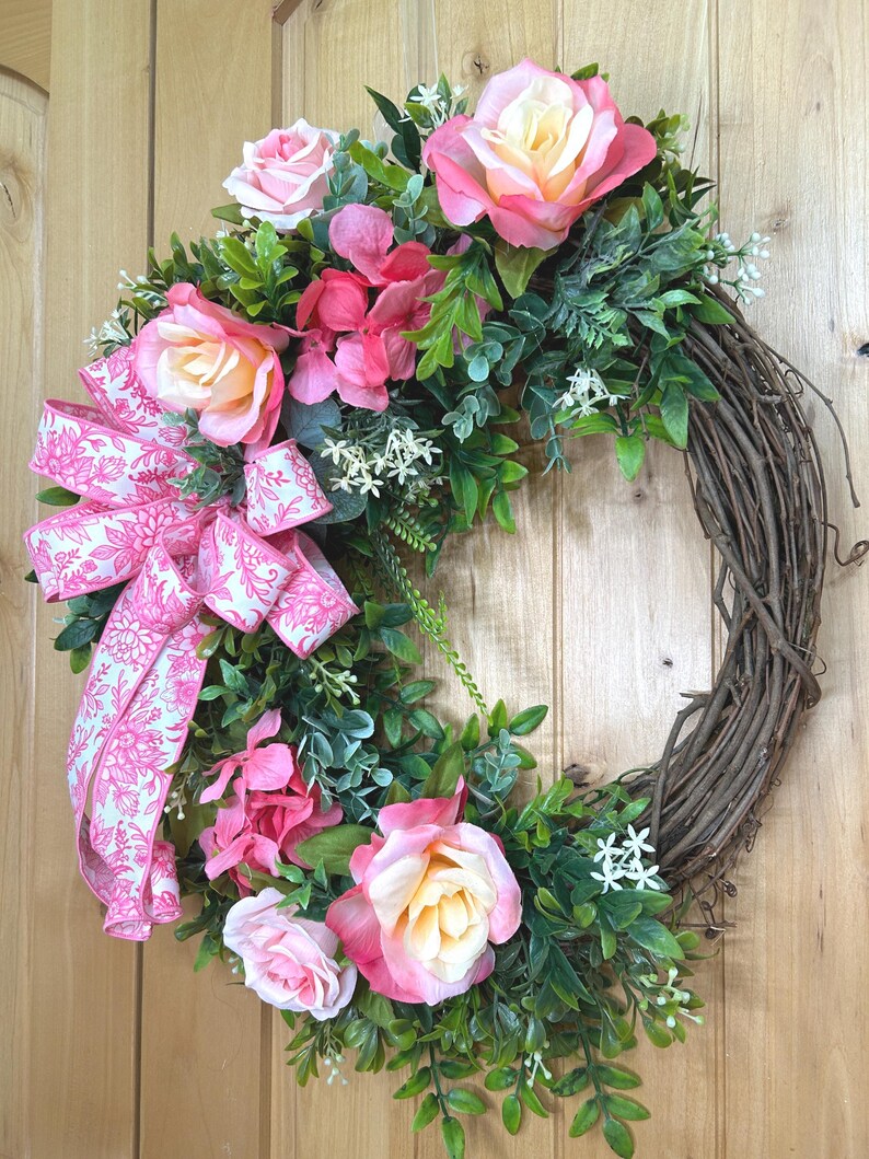 May include: A decorative wreath featuring a natural brown grapevine base adorned with artificial pink and cream roses, vibrant pink hydrangea clusters, and green foliage. A large, patterned pink and white floral ribbon bow is attached to the side.