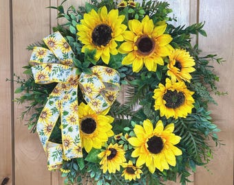 May include: A circular wreath featuring bright yellow sunflowers with dark brown centres, interspersed with lush green foliage and fern-like leaves. A decorative fabric ribbon with a repeating sunflower and green leaf pattern is tied to the left side.
