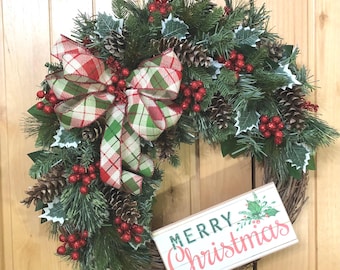 May include: A decorative wreath featuring green pine branches, brown pinecones, red berries, and white-tipped holly leaves. A large plaid ribbon in red, green, and white is tied at the top. A rectangular sign reads MERRY Christmas in red and green.