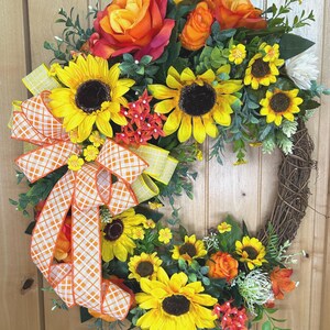 May include: A decorative wreath featuring artificial sunflowers and orange roses arranged on a brown twig base. The arrangement includes green foliage and small red floral accents, finished with a large orange and white checkered ribbon bow.