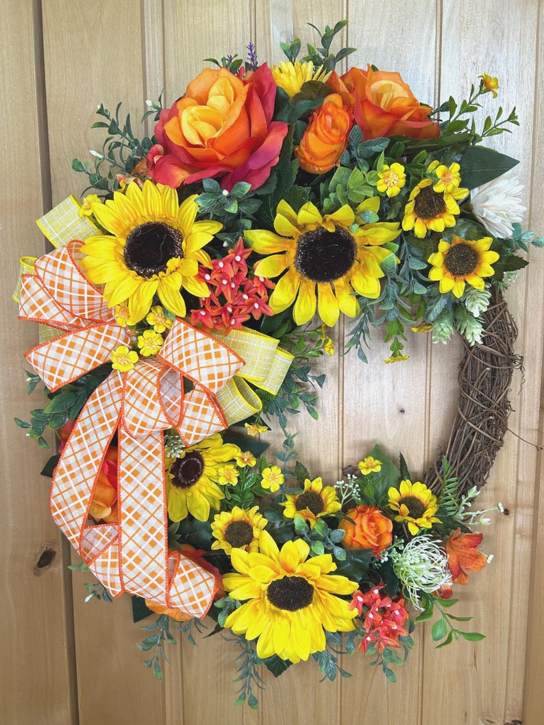 May include: A decorative wreath featuring artificial sunflowers and orange roses arranged on a brown twig base. The arrangement includes green foliage and small red floral accents, finished with a large orange and white checkered ribbon bow.