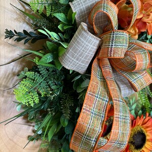 May include: A decorative wreath featuring artificial green foliage, ferns, and orange flowers. A large, multi-loop bow made of two different ribbons is attached to the center. One ribbon is a solid tan plaid, and the other is an orange, green, and white plaid.