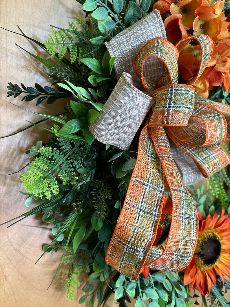May include: A decorative wreath featuring artificial green foliage, ferns, and orange flowers. A large, multi-loop bow made of two different ribbons is attached to the center. One ribbon is a solid tan plaid, and the other is an orange, green, and white plaid.