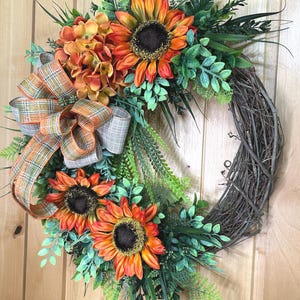 May include: A decorative wreath featuring three large orange and yellow sunflowers, a cluster of orange hydrangea-like flowers, and various green foliage attached to a circular brown twig base, accented with a plaid ribbon in orange, green, and tan.