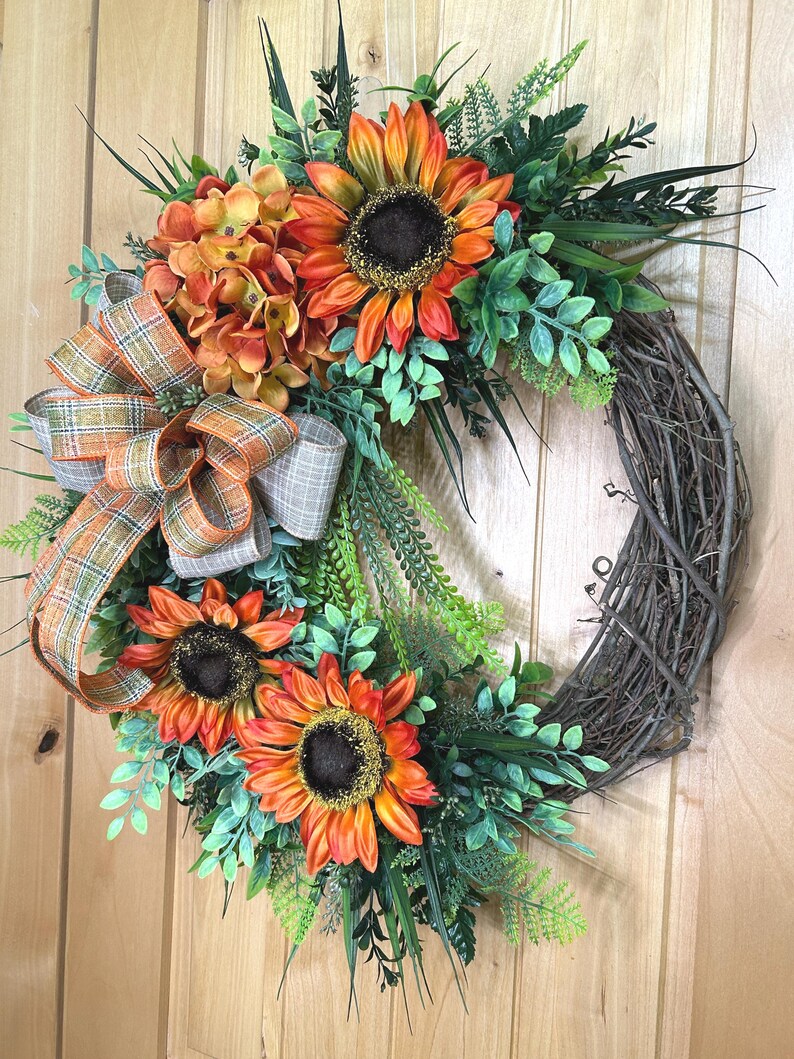 May include: A decorative wreath featuring three large orange and yellow sunflowers, a cluster of orange hydrangea-like flowers, and various green foliage attached to a circular brown twig base, accented with a plaid ribbon in orange, green, and tan.