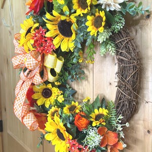 May include: A decorative wreath featuring artificial sunflowers, orange roses, and small white flowers arranged on a brown twig base. An orange and white checkered ribbon is tied to the side, accented with green foliage and small filler plants.