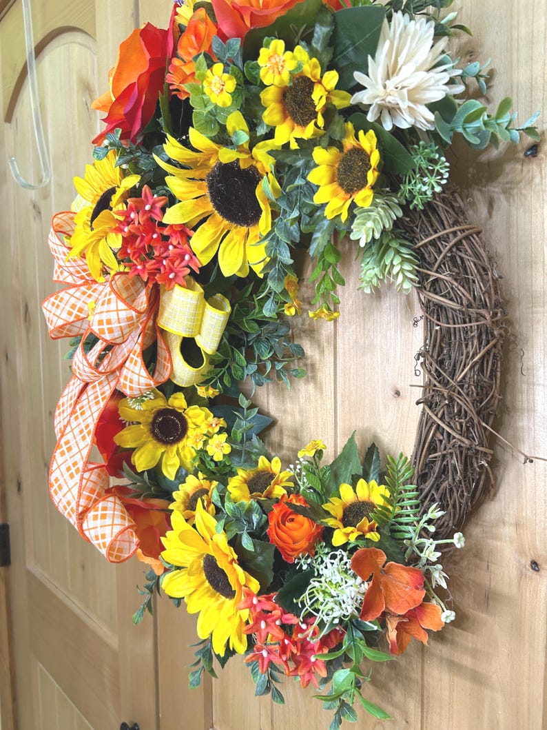 May include: A decorative wreath featuring artificial sunflowers, orange roses, and small white flowers arranged on a brown twig base. An orange and white checkered ribbon is tied to the side, accented with green foliage and small filler plants.