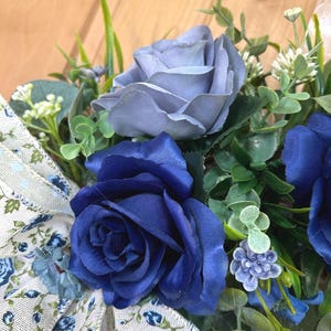 Op de afbeelding: Een bloemstuk met diep marineblauwe en stoffig blauwe rozen, geaccentueerd met groen blad, kleine witte bloemetjes en een beige lint met een blauw bloemenpatroon. De compositie bevat diverse bladeren en stengels tussen de rozen.